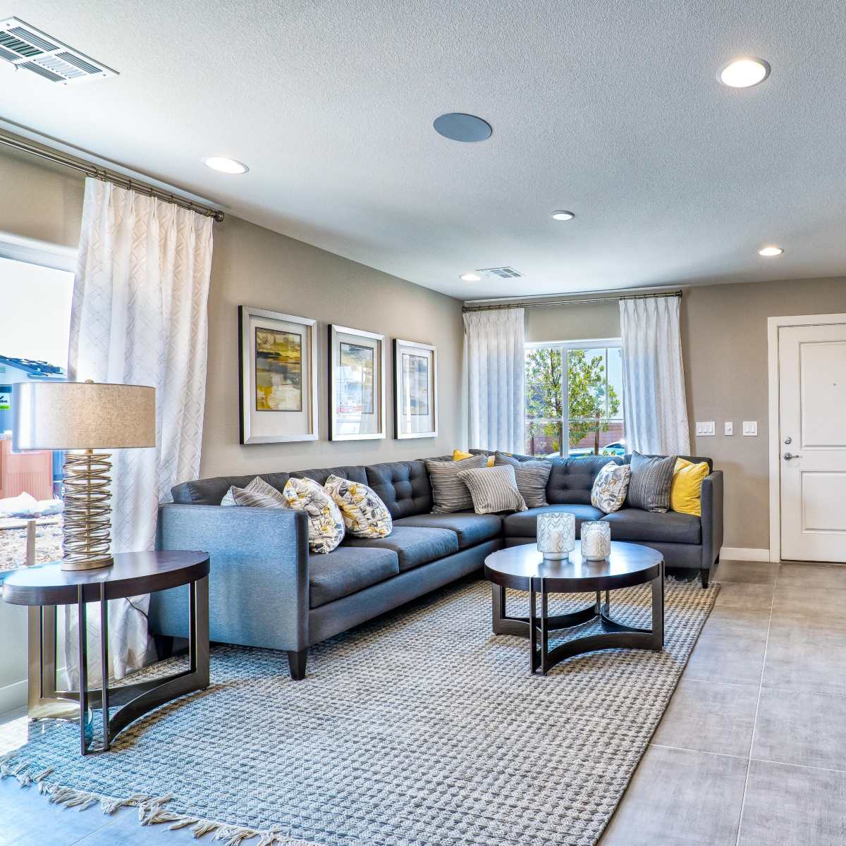 A modern living room features a sectional sofa, a coffee table, and a television mounted on the wall, with windows allowing natural light to enter.