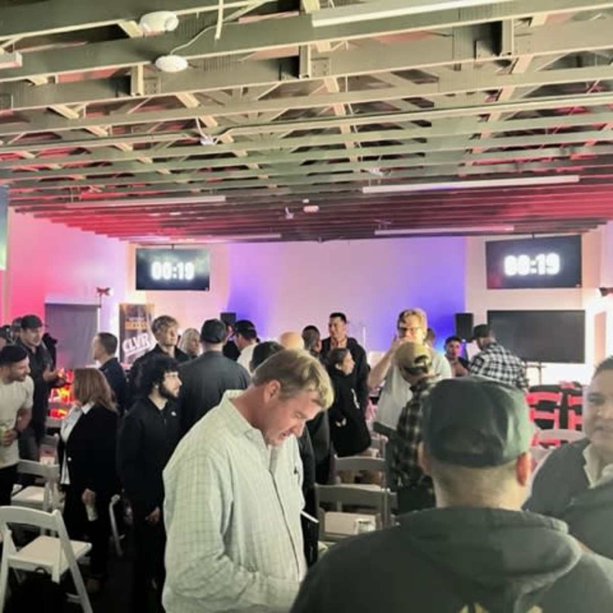 A crowded event space features large digital countdown timers on the walls, with attendees mingling and sitting in white chairs.