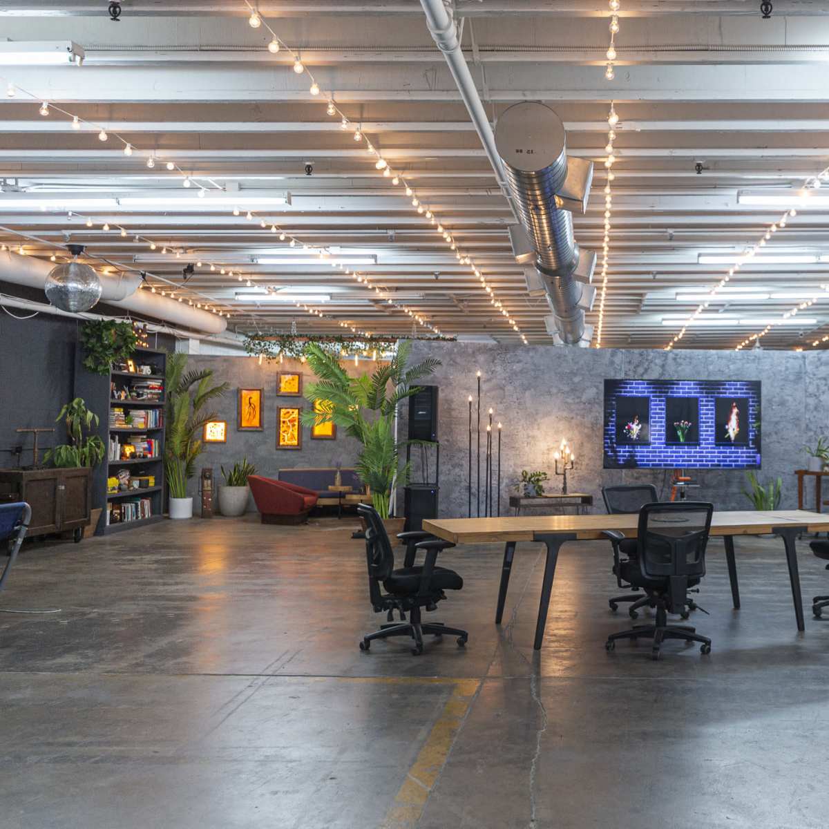 A spacious, industrial-style meeting room features a long wooden table surrounded by black office chairs, with a large screen displaying information on a gray textured wall.