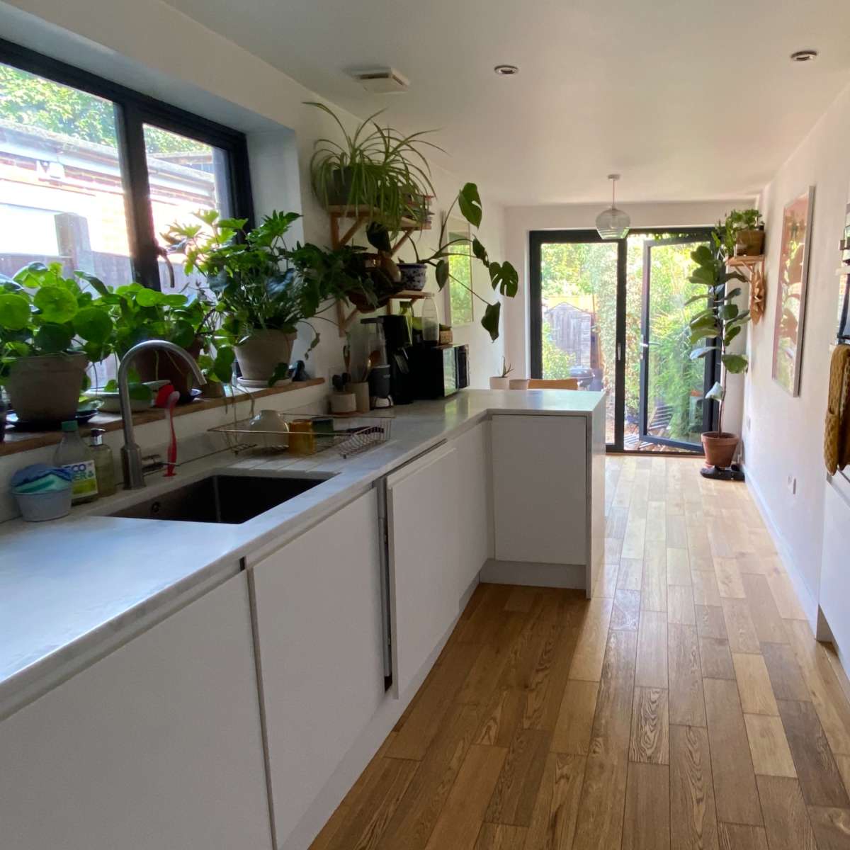 A modern kitchen with white cabinetry, wooden flooring, and large windows facing a garden filled with plants.