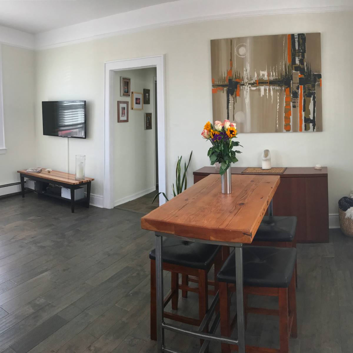 A dining area features a wooden table with black chairs, surrounded by minimalist decor, a painting on the wall, and a bouquet of flowers in a vase.