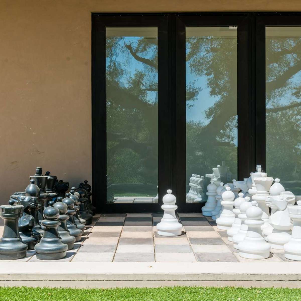 A large outdoor chessboard displays oversized black and white chess pieces arranged for a game in front of glass doors.