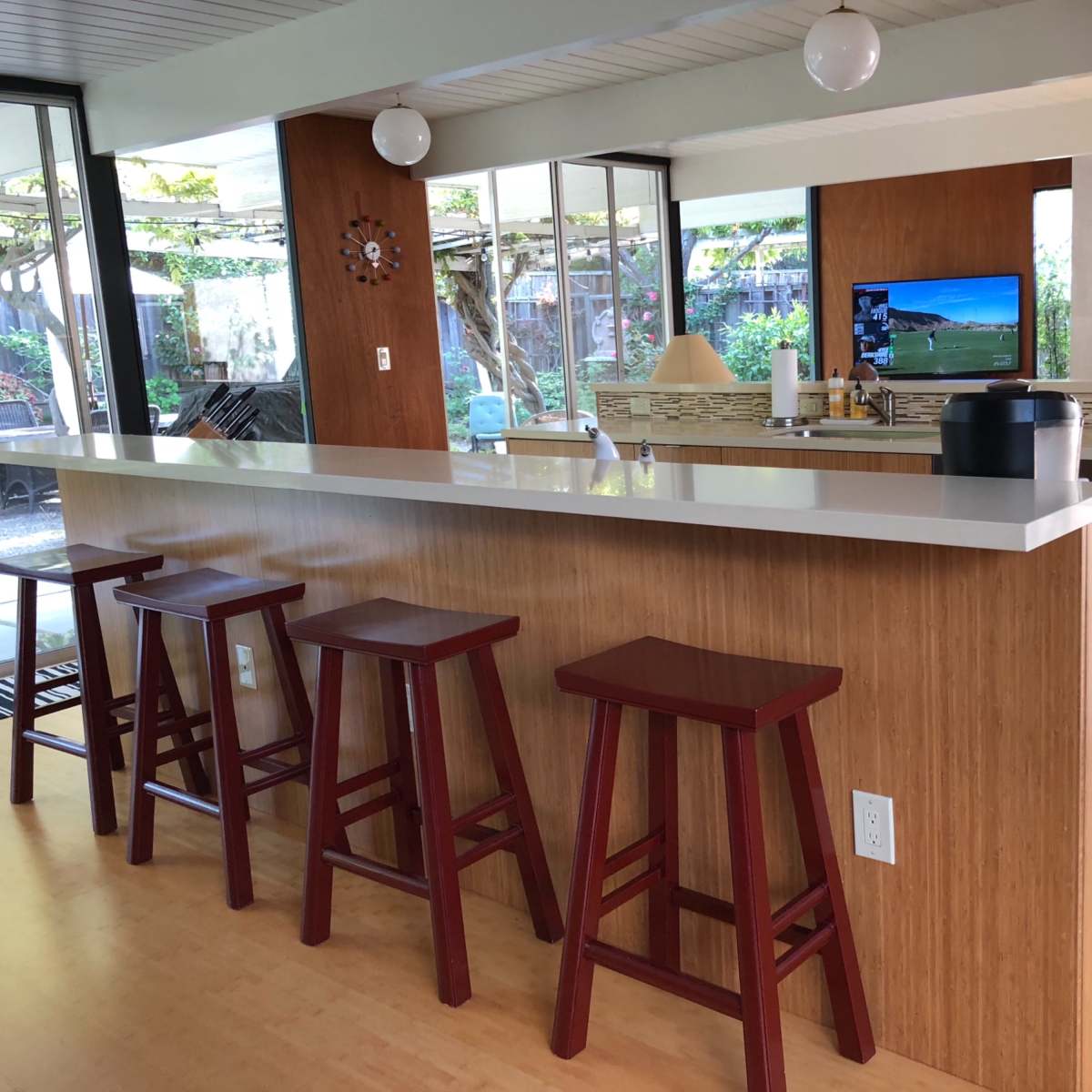 A modern kitchen with a bar counter and four wooden stools, featuring a television and windows that overlook a garden area.