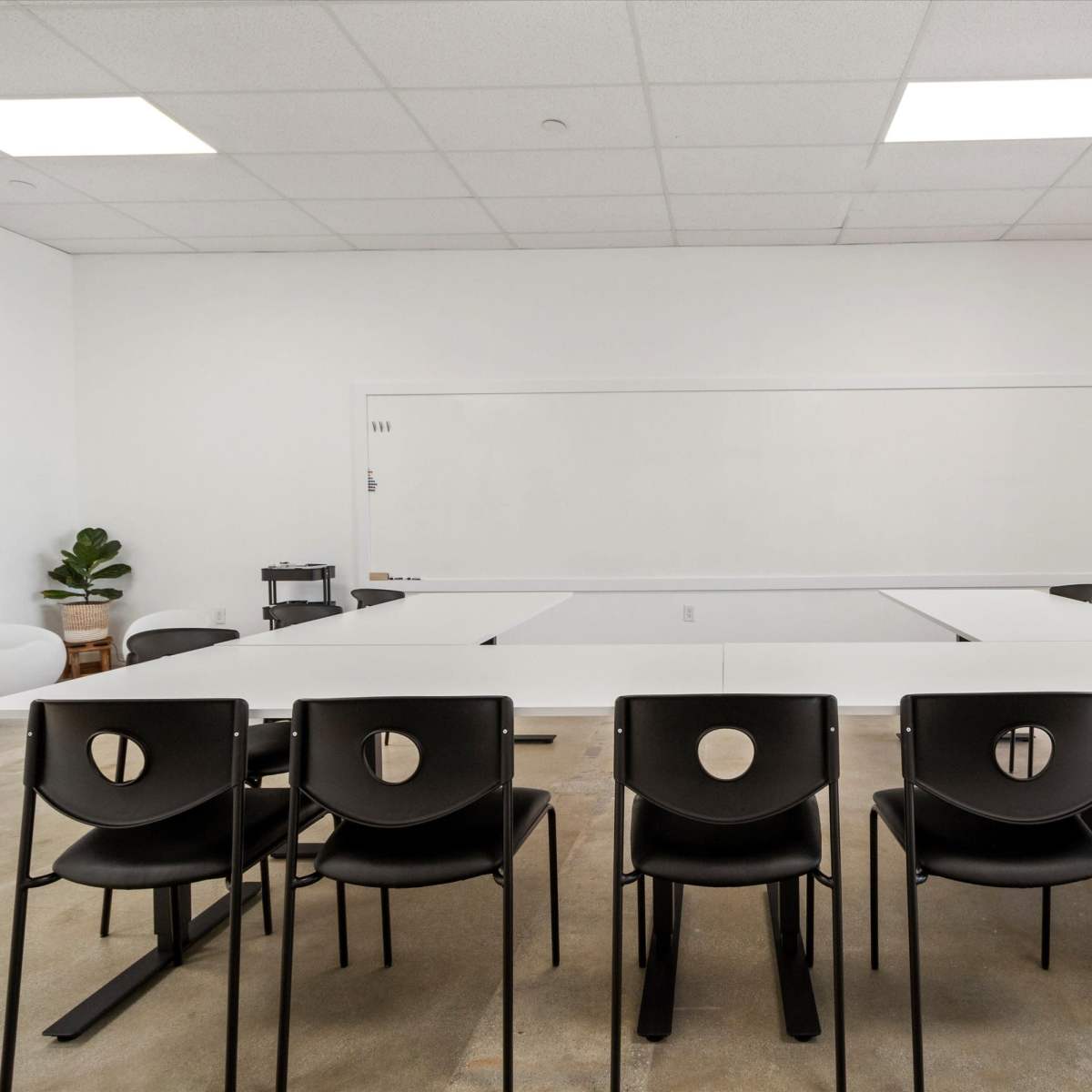 A large, minimalist meeting room features a rectangular table surrounded by black chairs, with a whiteboard and plants in the background.