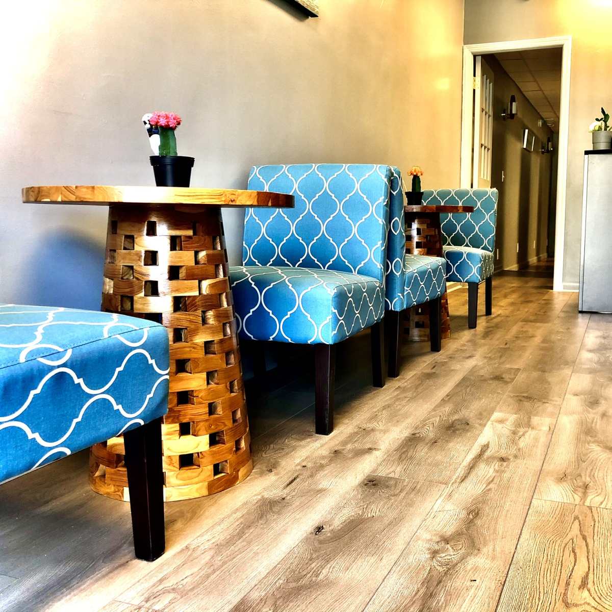 A line of blue chairs with a geometric pattern and wooden tables, set in a narrow hallway with light-colored flooring.