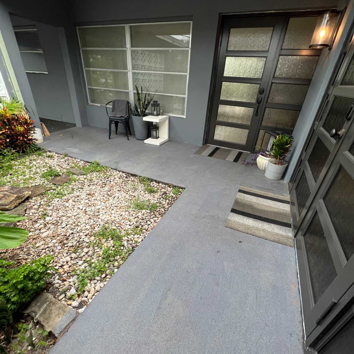 A residential entryway with a paved area leading to a glass-paneled front door, flanked by plants and gravel landscaping.