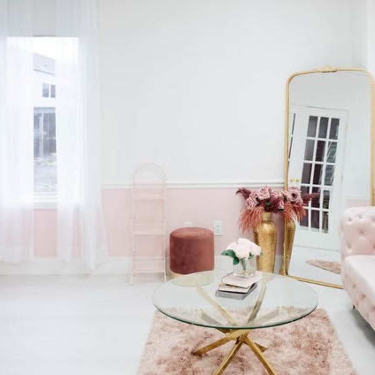 The room features a light-colored décor with a pink tufted sofa, a glass coffee table, a large mirror, and decorative plants.