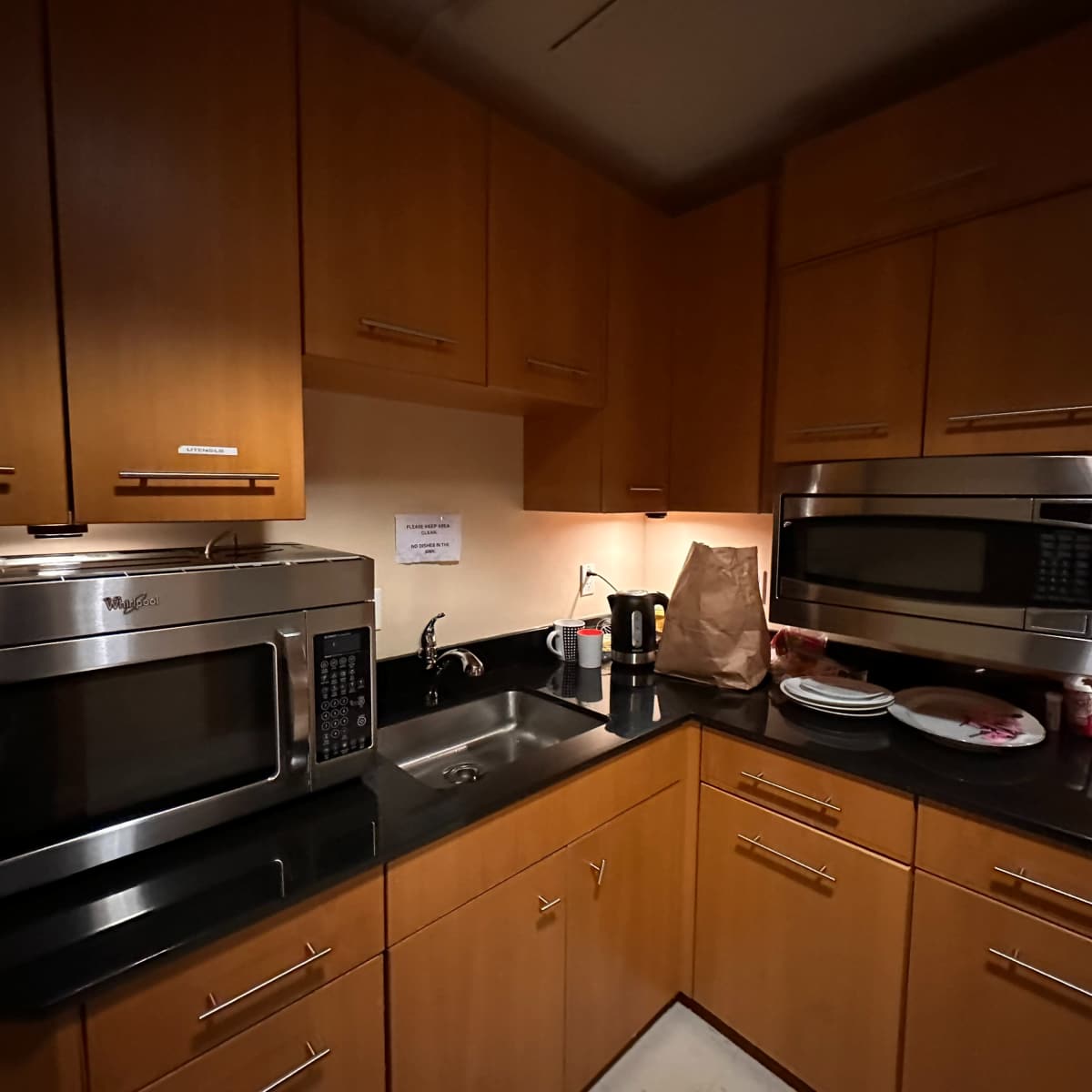 A compact kitchen with wooden cabinets, a stainless steel microwave and a coffee maker, and a sink beside a stack of plates.