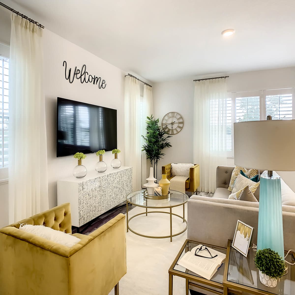 A modern living room with a light-colored sofa, a glass coffee table, and a television mounted on a white wall, decorated with a "Welcome" sign and plants.