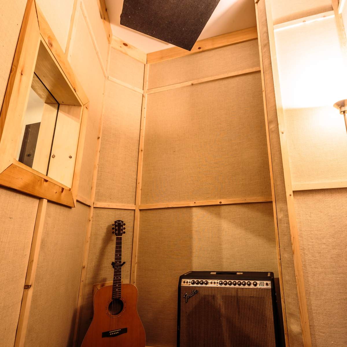 A soundproof recording studio booth featuring a guitar leaning against the wall, an amplifier, a lamp, and acoustic panels on the walls.