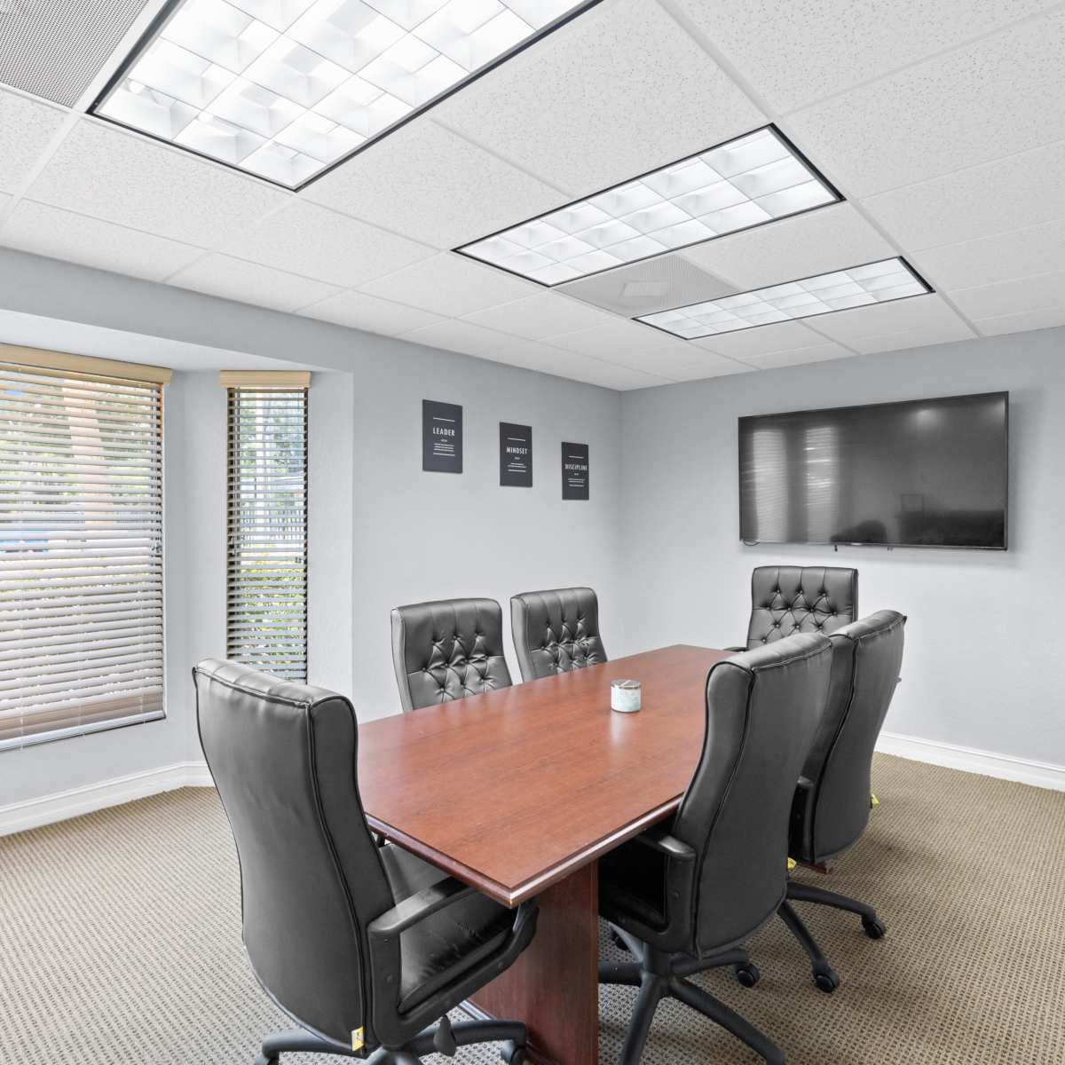 A modern conference room with a rectangular wooden table surrounded by black leather chairs, a wall-mounted TV, and large windows with blinds.