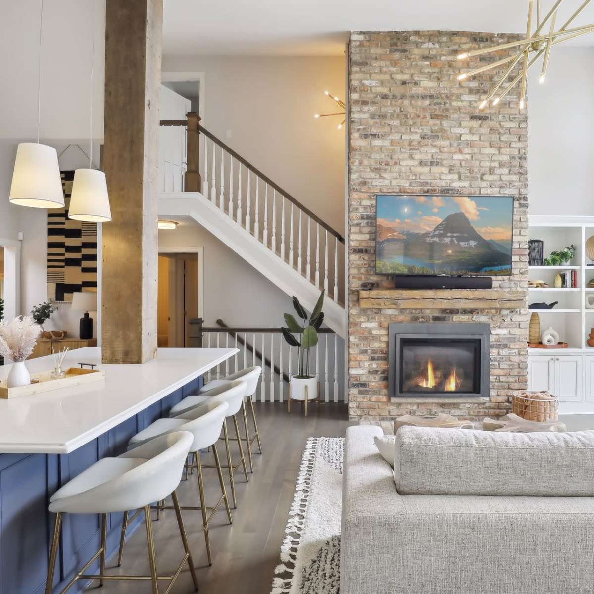 A modern living area with a stone fireplace, a large sectional sofa, and a staircase leading to an upper level, all against a backdrop of large windows overlooking a body of water.