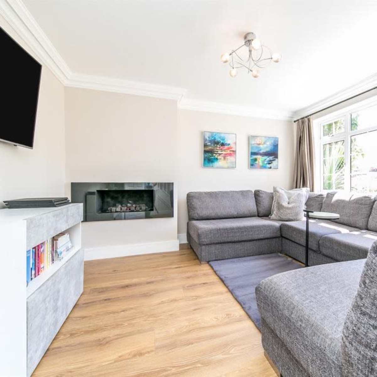 A modern living room with a sectional gray sofa, a wall-mounted television, a sleek electric fireplace, and large windows allowing natural light to enter.