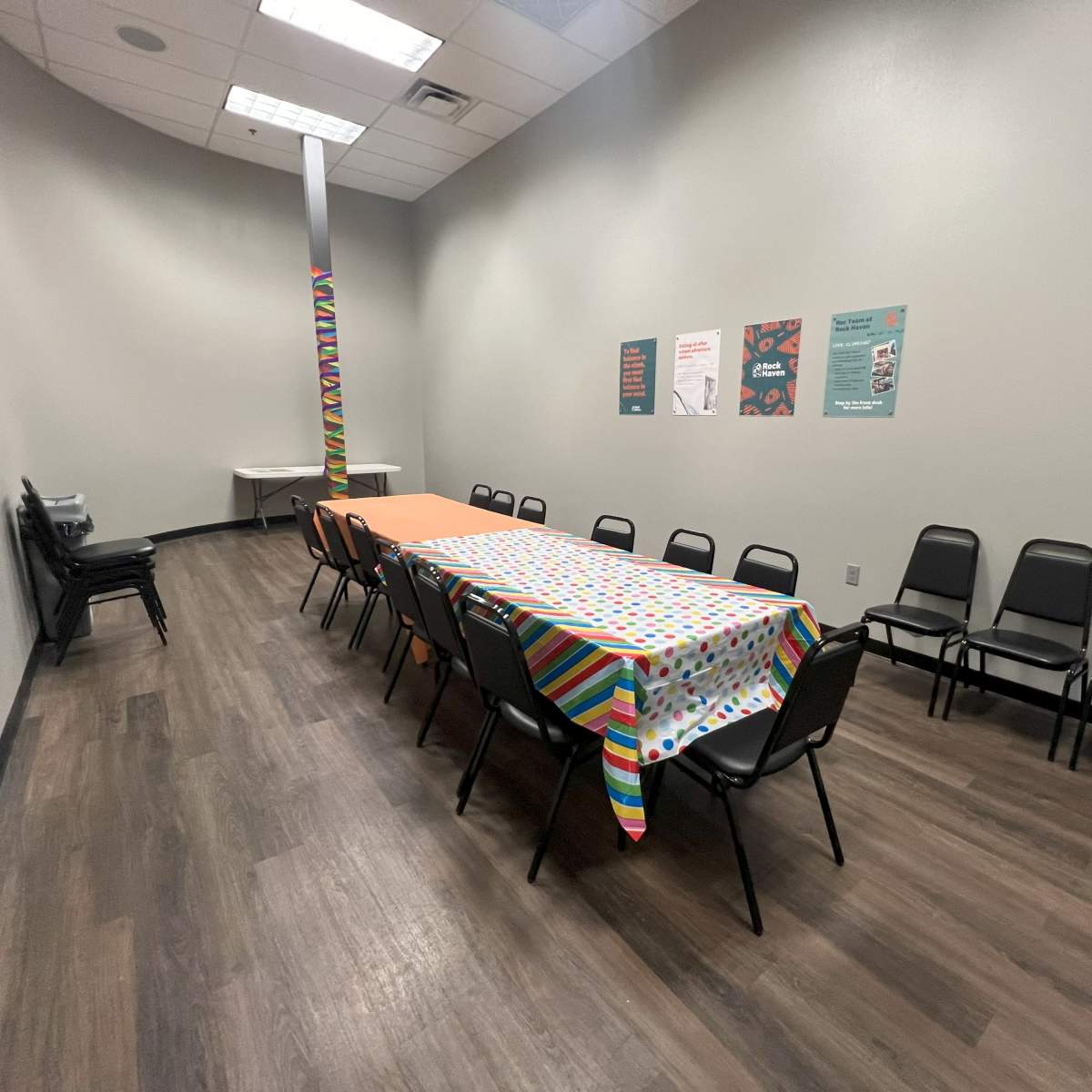 A long table covered with a patterned tablecloth is set up in a room with gray walls and black chairs arranged around it.