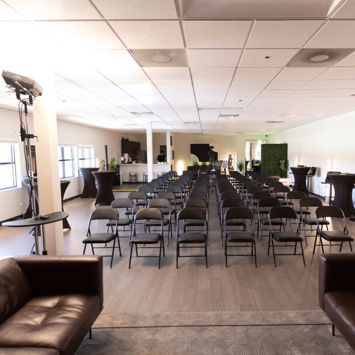 A spacious room arranged with rows of black chairs facing a presentation area, flanked by a bar and two leather couches.