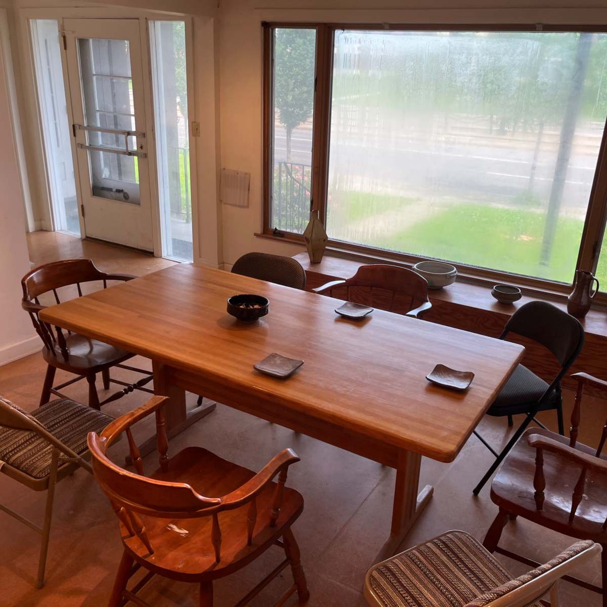 A wooden dining table is surrounded by six chairs in a well-lit room with large windows overlooking a street.