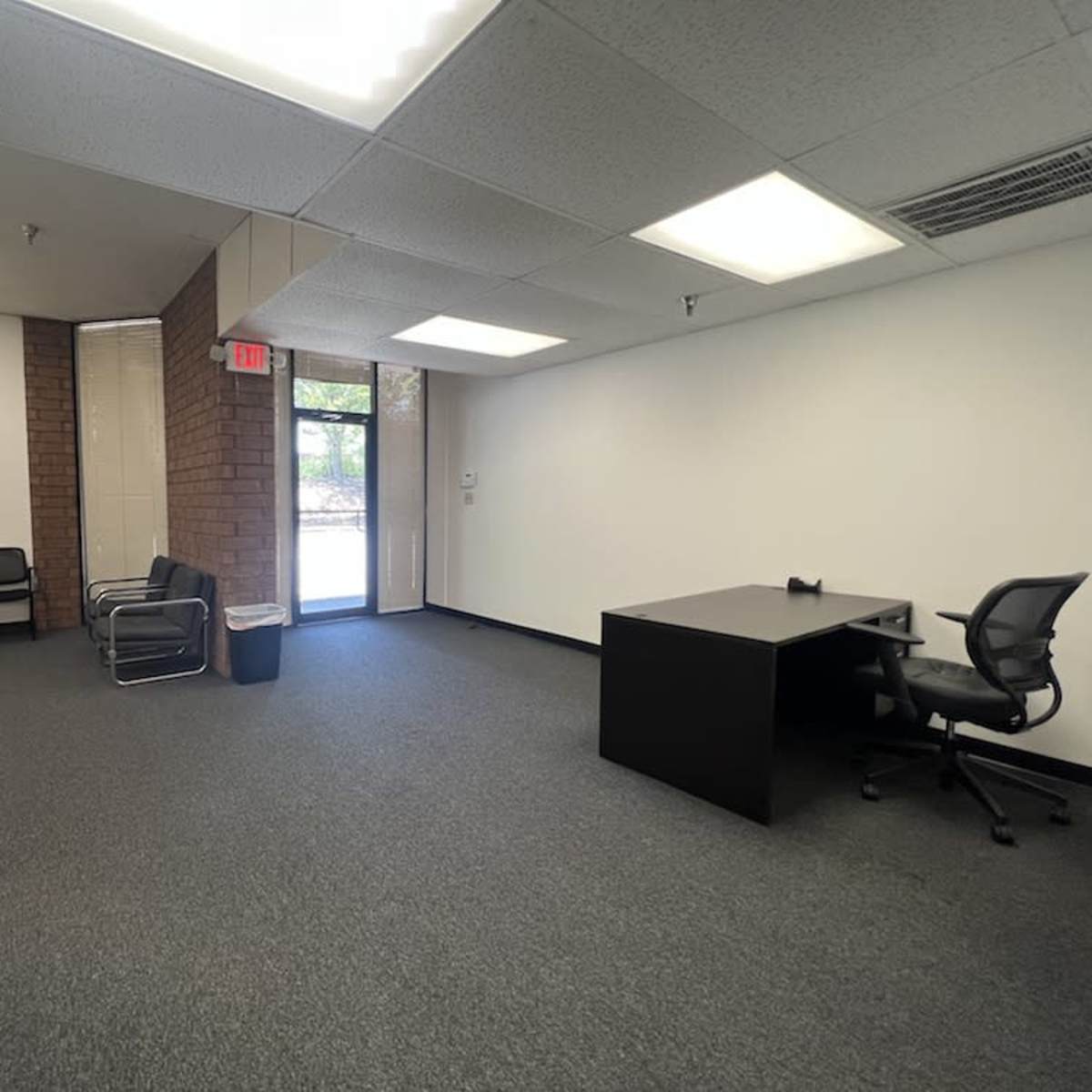 A partially furnished office space with a few chairs along one wall, a desk and chair, and large windows letting in natural light.