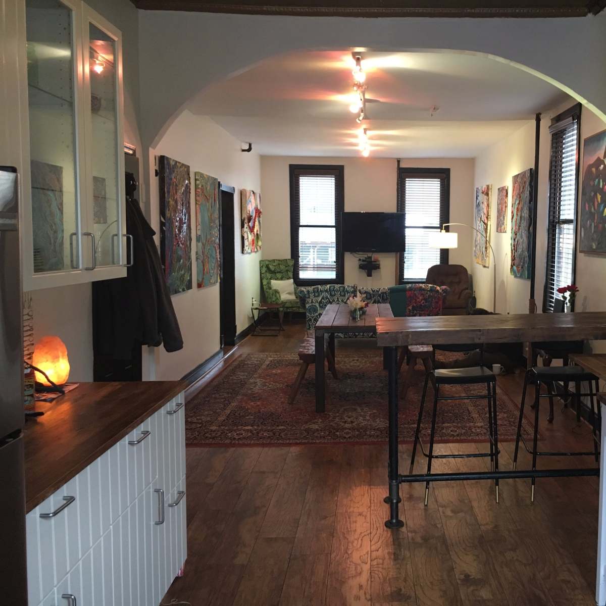 A spacious, well-lit living area with wooden floors, art on the walls, and a mix of seating options, including a couch and dining table.