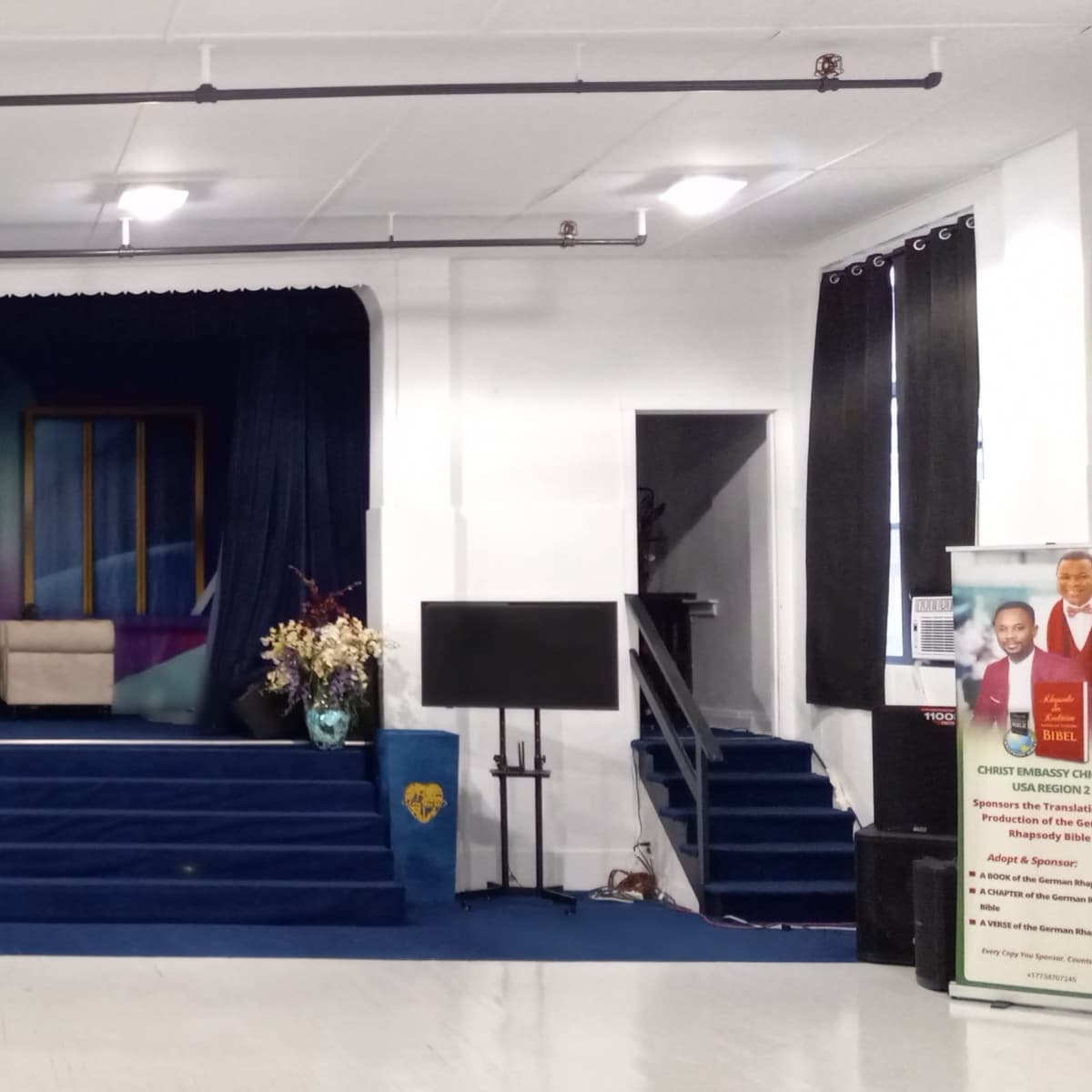 A stage with blue curtains, a central seating area, and two large monitors on either side, set in a spacious, well-lit room.