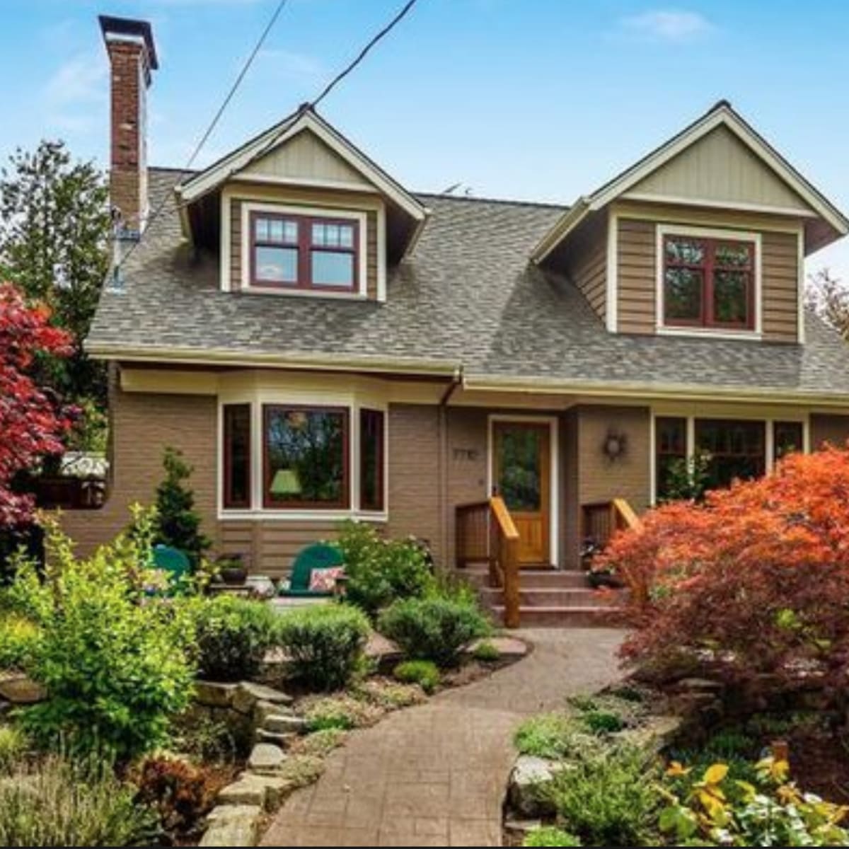 A two-story house with a sloped roof, surrounded by a landscaped garden featuring colorful shrubs and a stone pathway.