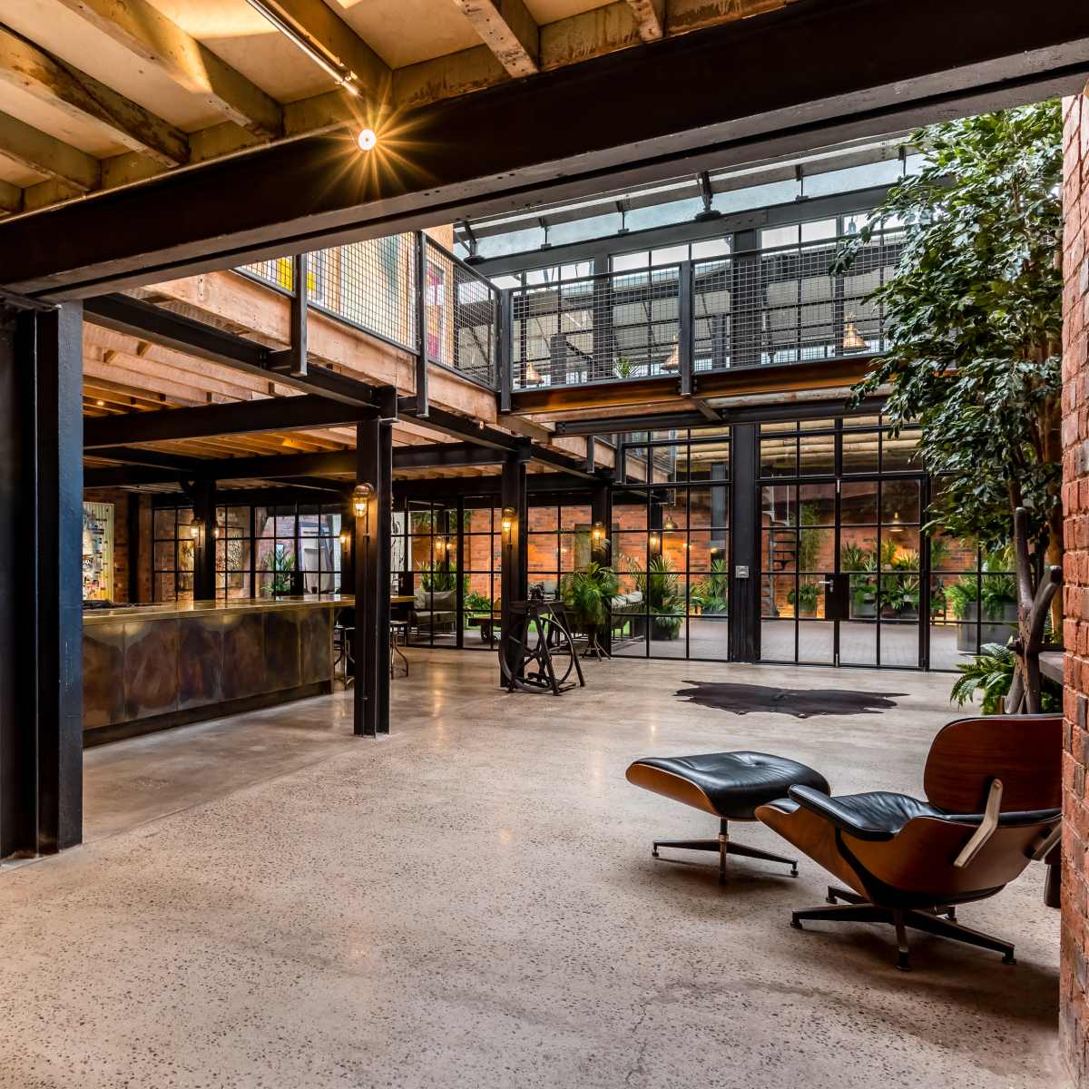 A spacious industrial-style interior with exposed brick walls, large windows, and a mix of modern furniture and plants.