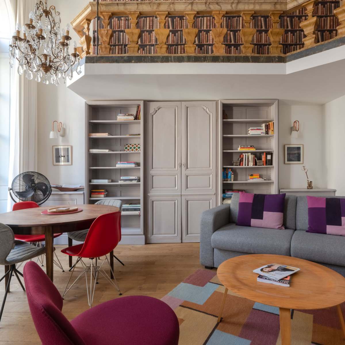 A cozy living room with a gray sofa, a circular wooden table surrounded by red and purple chairs, and a bookshelf on the wall.
