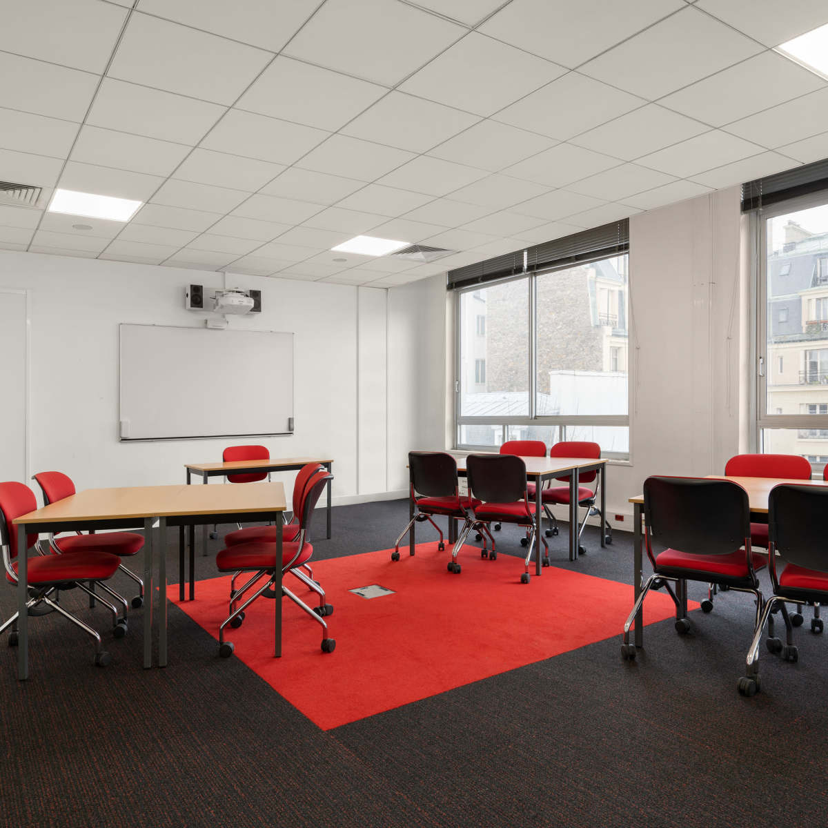 A modern meeting room with red chairs around several tables and large windows overlooking a cityscape.