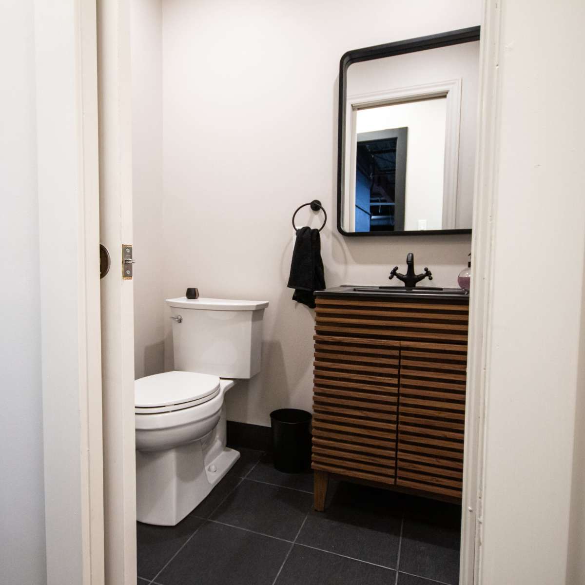 A small bathroom features a toilet, a dark wooden vanity with a sink, and a mirror above it, all illuminated by natural light.