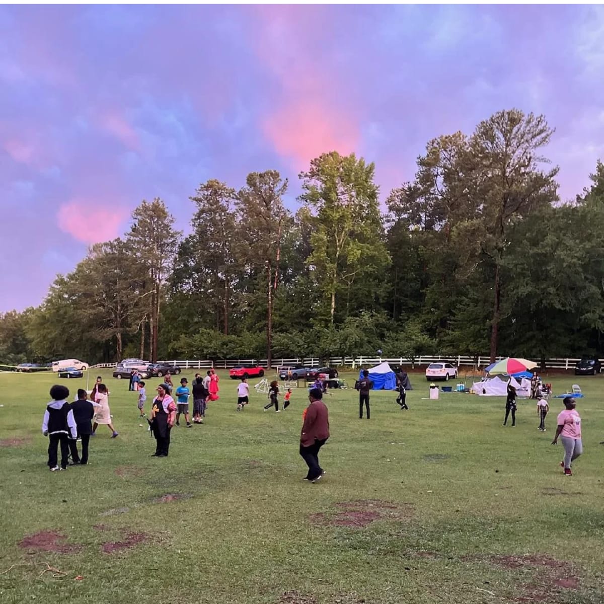 A group of people are gathered on a grassy field, some engaging in activities while others are scattered throughout the area, with cars and tents visible in the background.