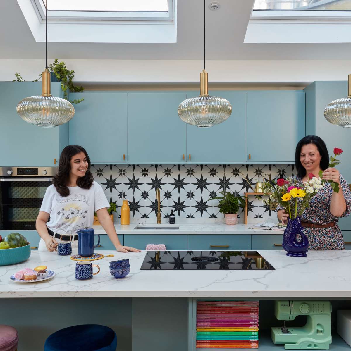 Two women are arranging flowers in a modern kitchen with blue cabinets and patterned tiles, while items like fruit and dishes are displayed on the island.