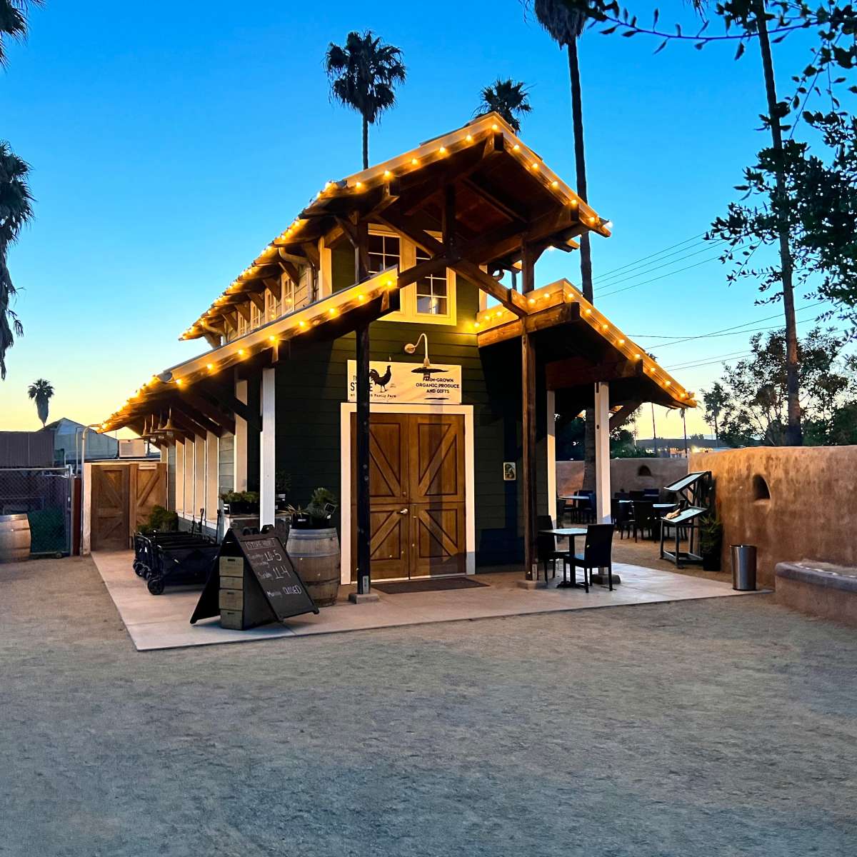 A two-story building with a modern rustic design, featuring outdoor seating and decorative string lights, surrounded by palm trees at dusk.