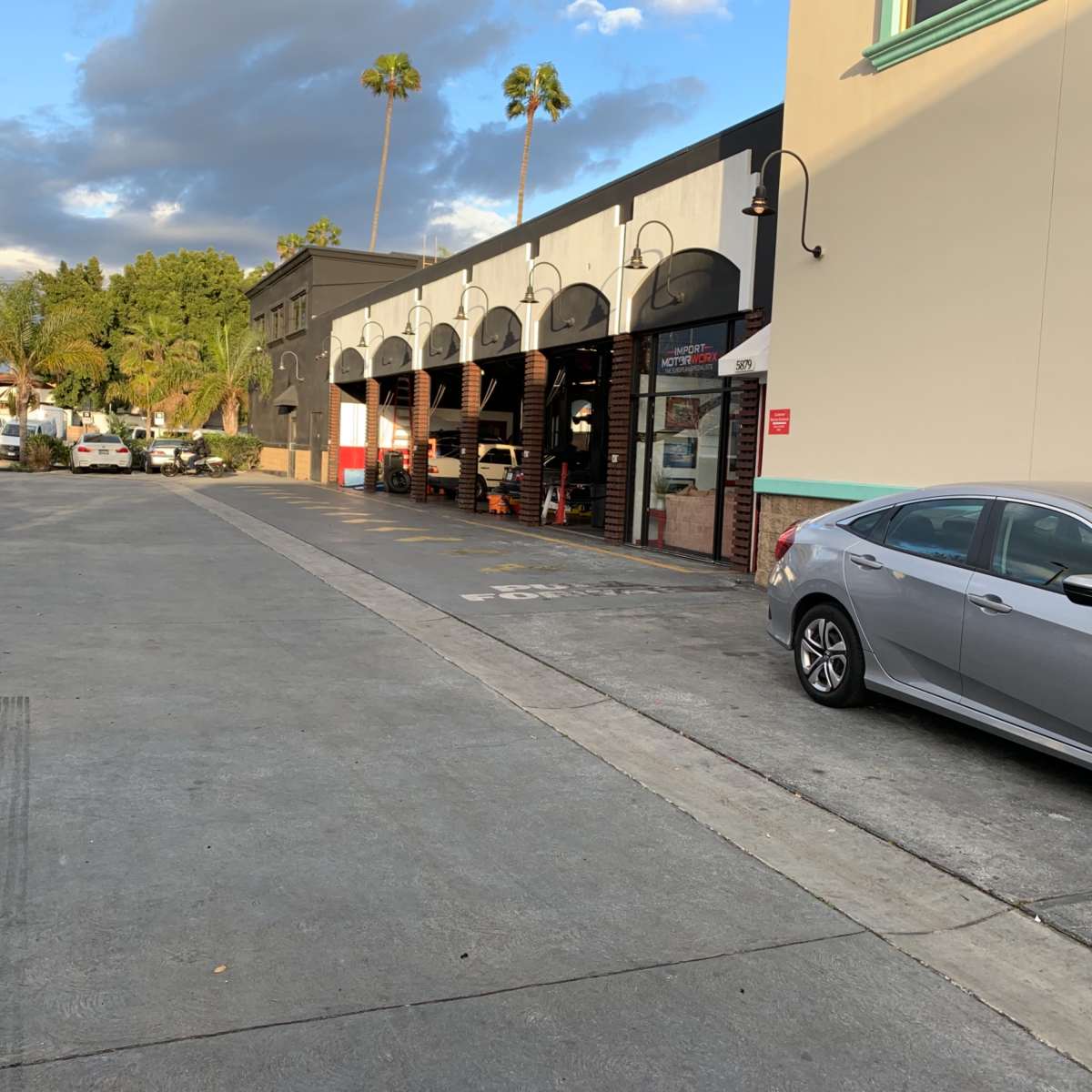 A row of buildings with arched openings lines a paved street, accompanied by parked cars and palm trees in the background.