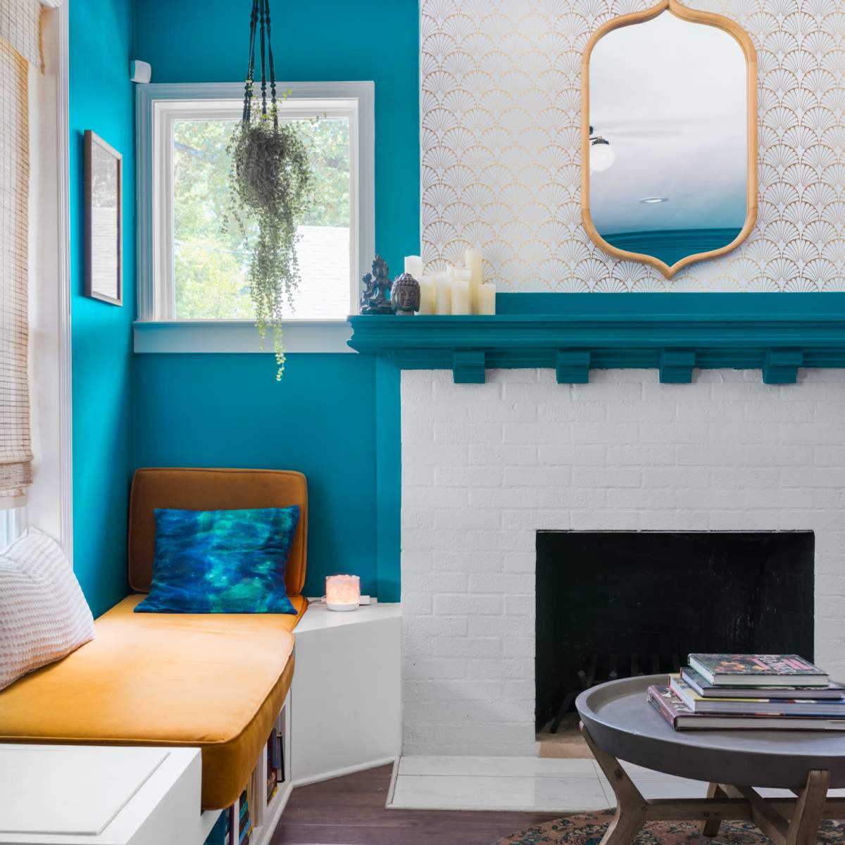 A bright room with turquoise walls, featuring a cozy seating area with an orange cushion, a white brick fireplace, and a round coffee table stacked with books.