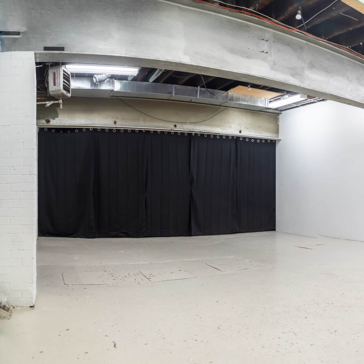 A spacious, empty room with exposed beams and a concrete floor, featuring black curtains on one wall and a white wall adjacent to a garage door.