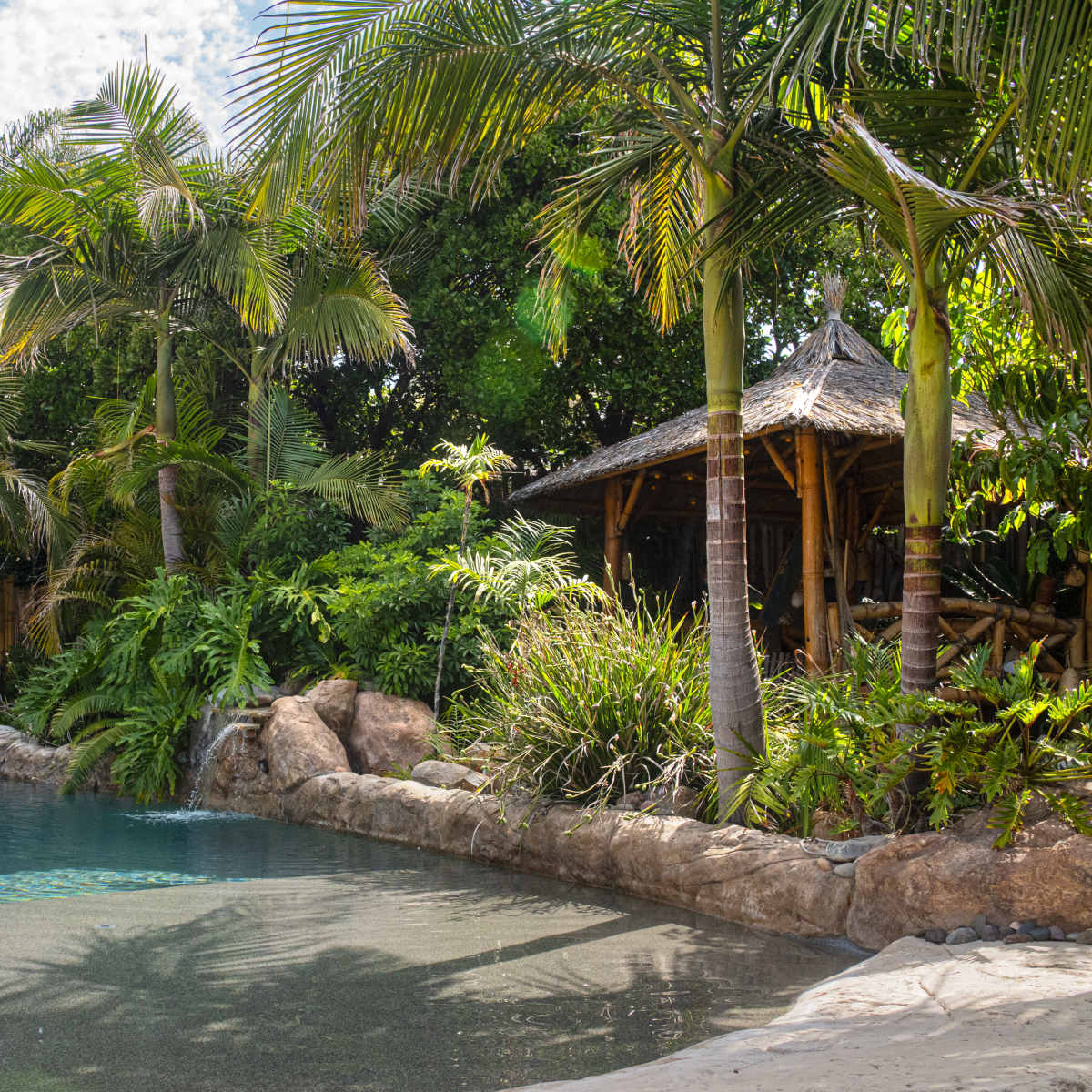 A tranquil pool is surrounded by tropical plants and rocks, with a thatched-roof gazebo in the background.
