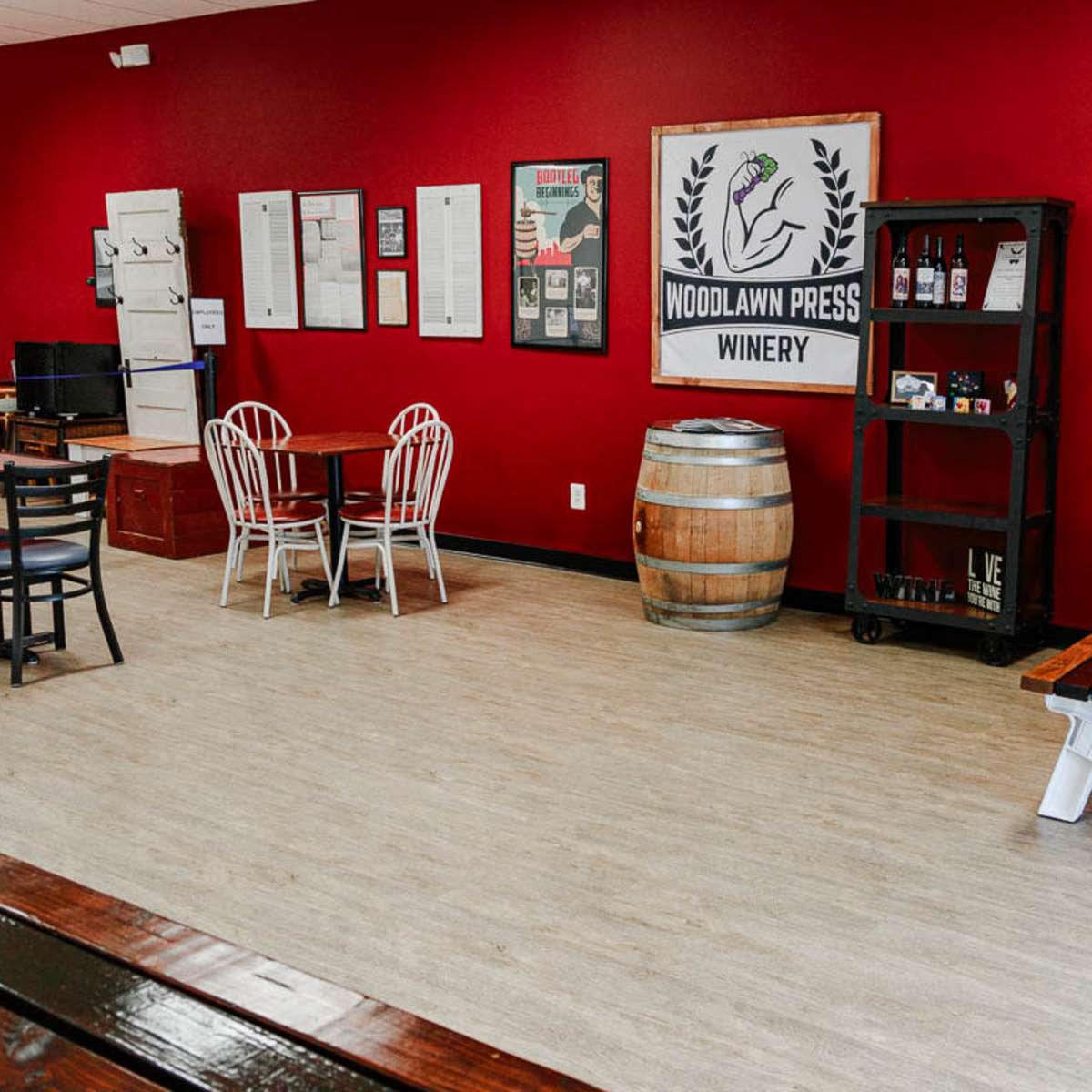 The interior of a winery features red walls, wooden tables, chairs, and a barrel, with various signage and decor on display.