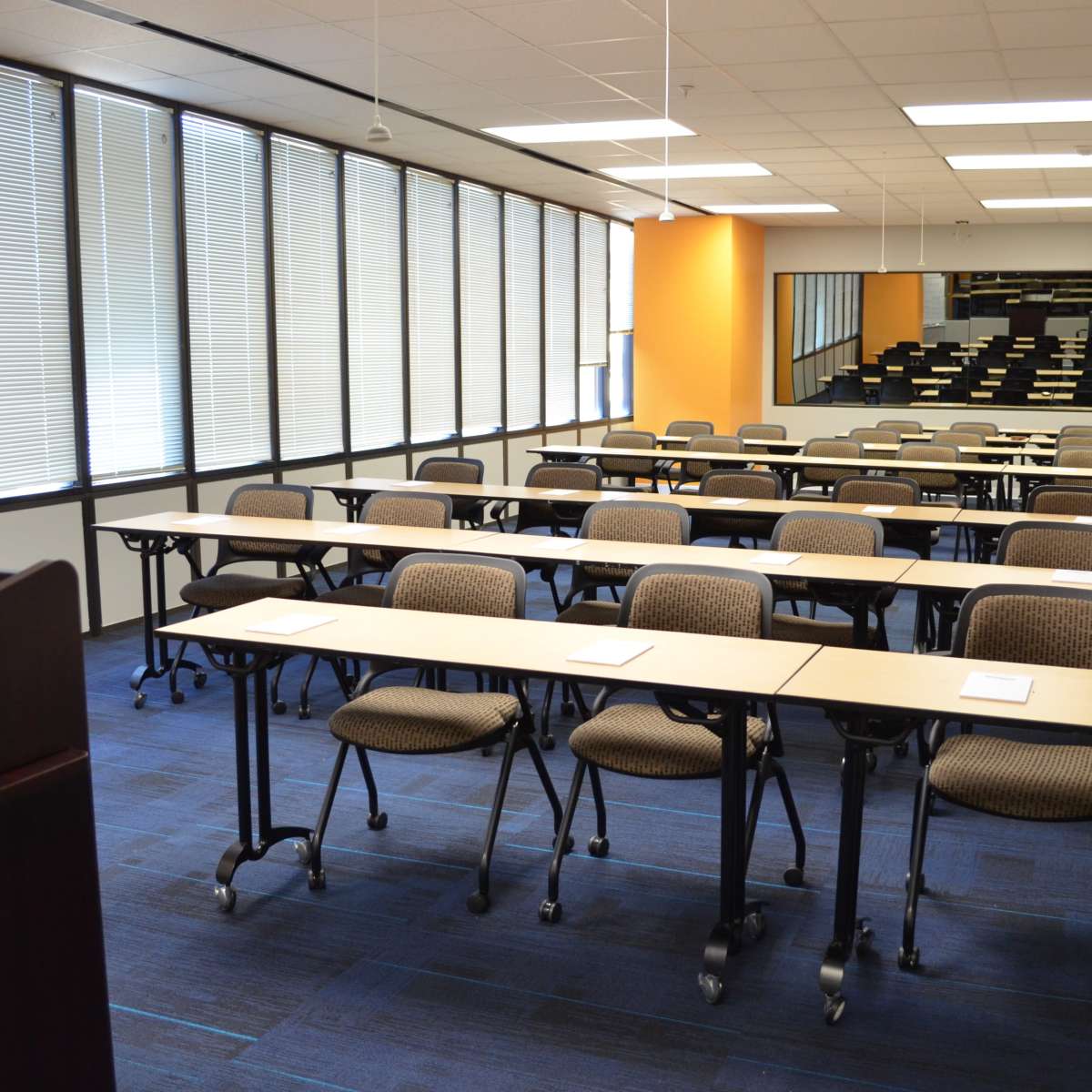 A classroom with rows of tables and chairs facing a presentation area, featuring large windows on one side and a staircase leading to an elevated section.