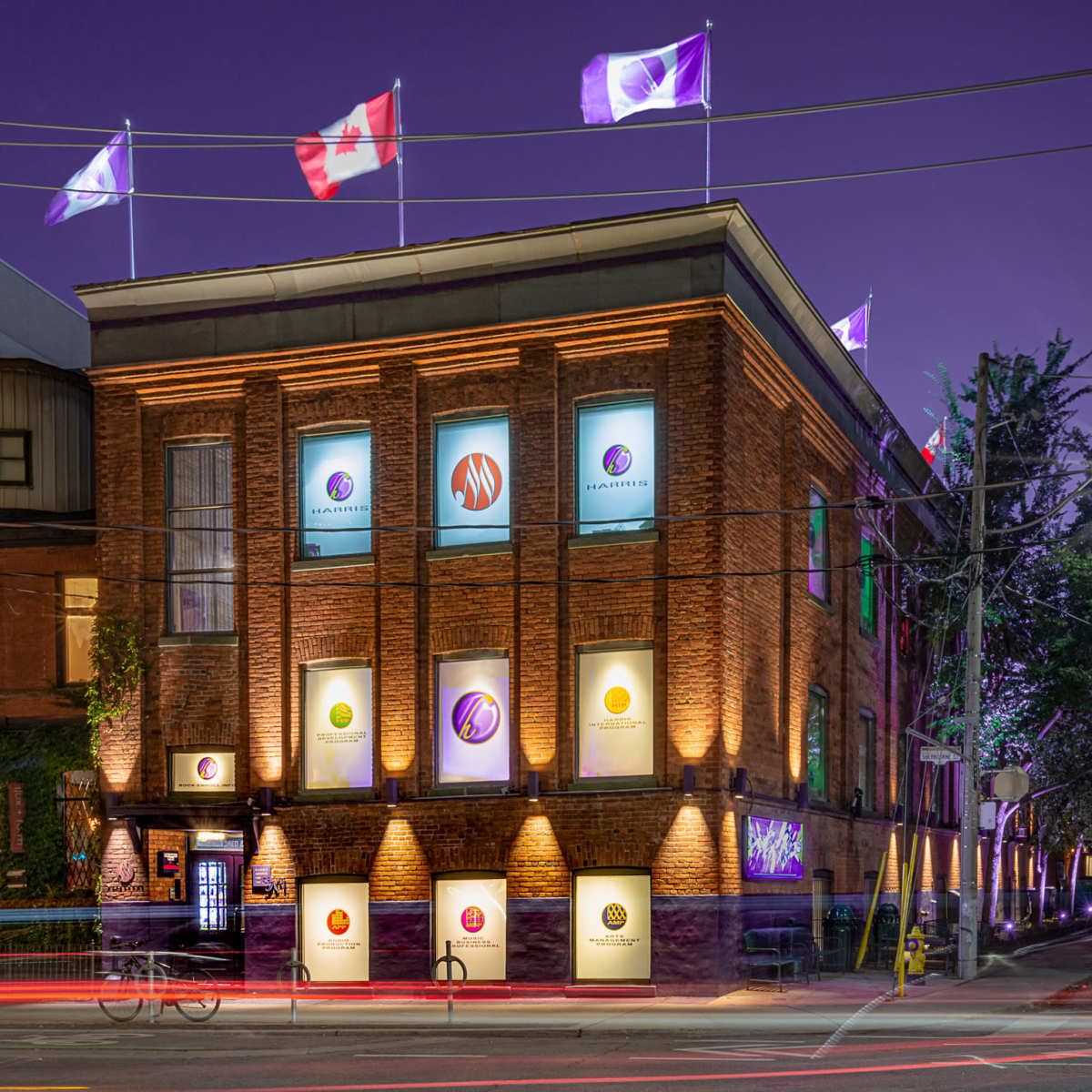 A brick building displays illuminated windows and several flags, including the Canadian flag, against a twilight sky.