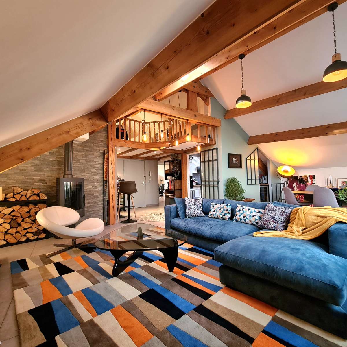 A well-furnished living room with a large blue sectional sofa, a coffee table, a colorful area rug, and exposed wooden beams on the ceiling.