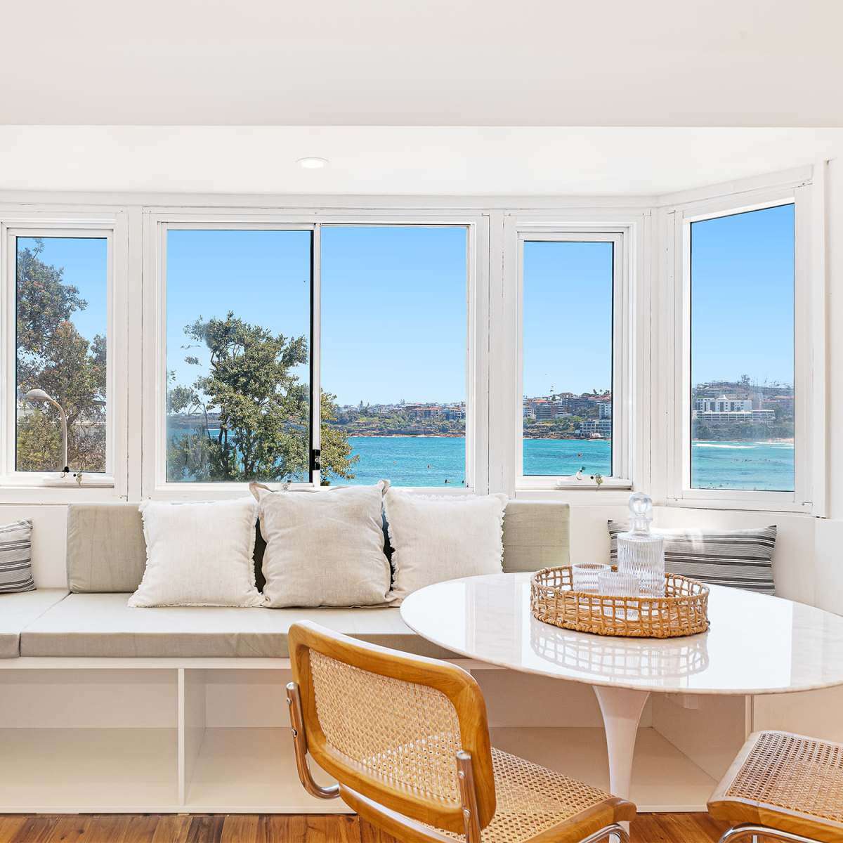 A bright, modern seating area features a bay window with views of a beach and sea, complemented by a round dining table and stylish chairs.