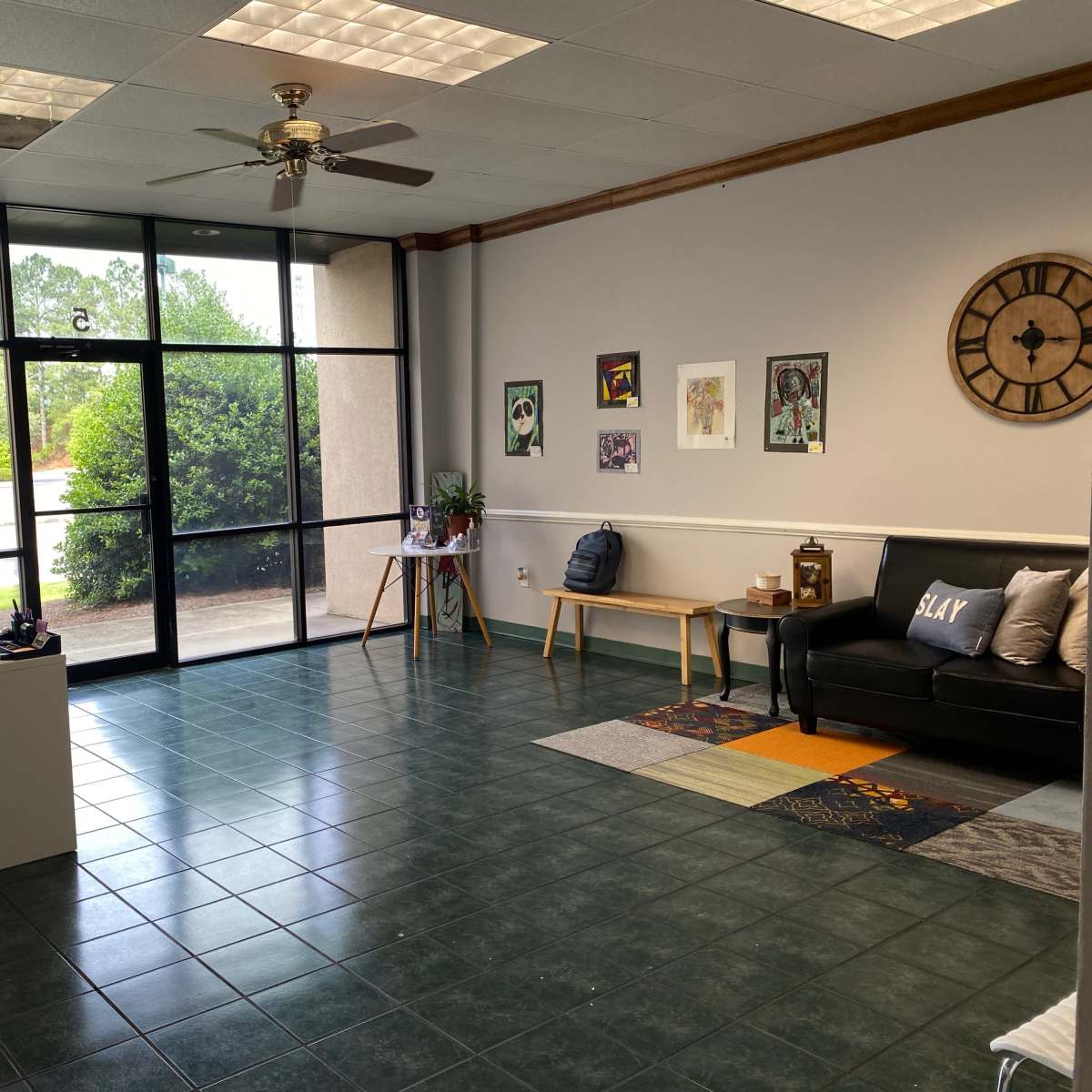 An empty office space with a black sofa, a small desk, and large windows allowing natural light to enter.