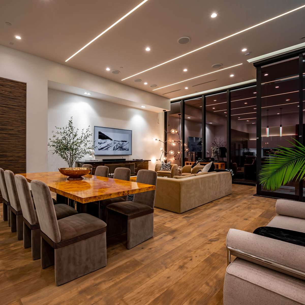 A modern living space features a dining table surrounded by upholstered chairs, a sofa, and large glass windows with a view of outdoor greenery.