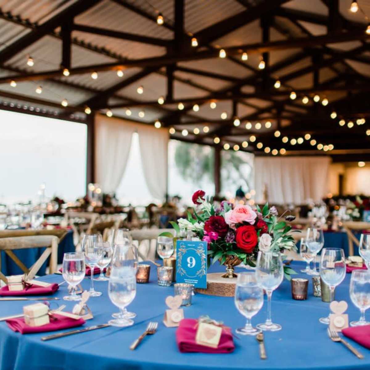 A banquet hall features elegantly set tables with blue tablecloths, floral centerpieces, and string lights overhead.