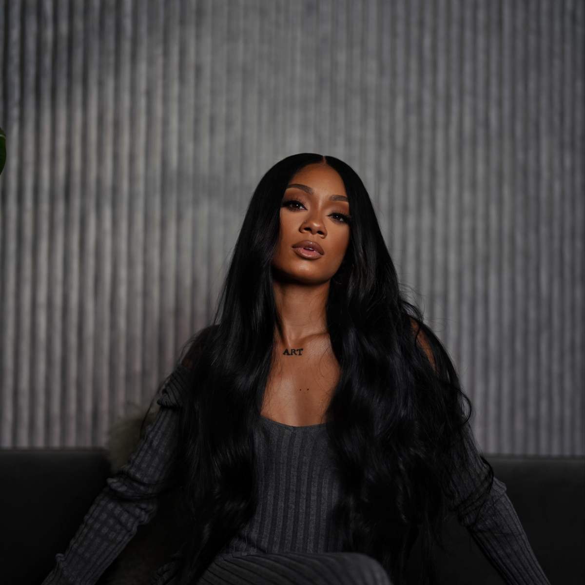 A person with long black hair sits on a dark sofa against a textured gray wall, wearing a form-fitting gray outfit.