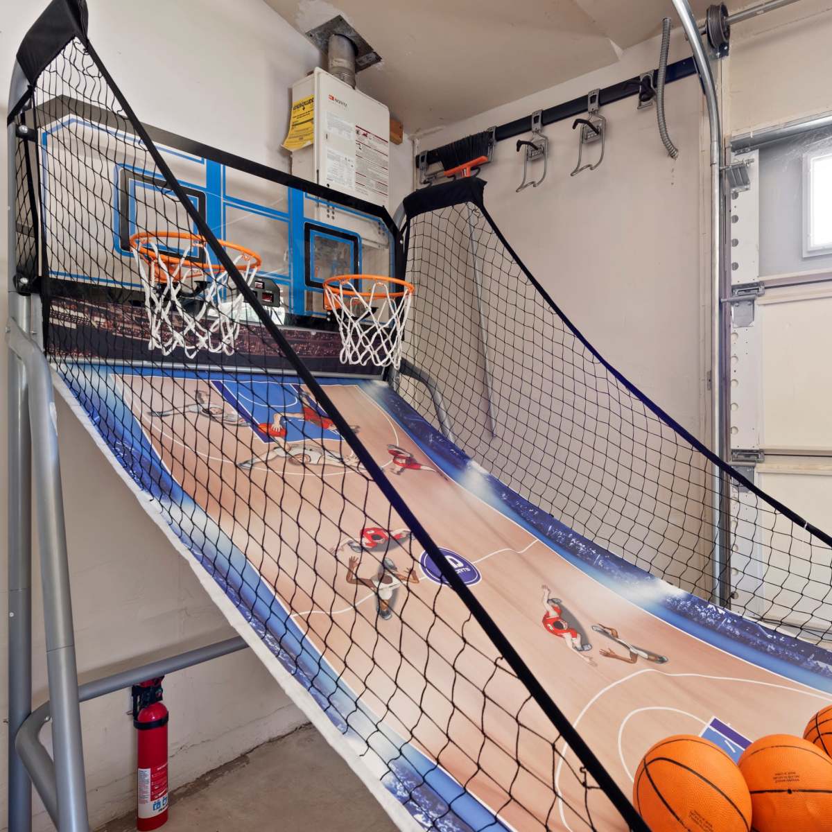 A home basketball arcade game set up in a garage with two basketball hoops, a sloped playing surface, and several orange basketballs.