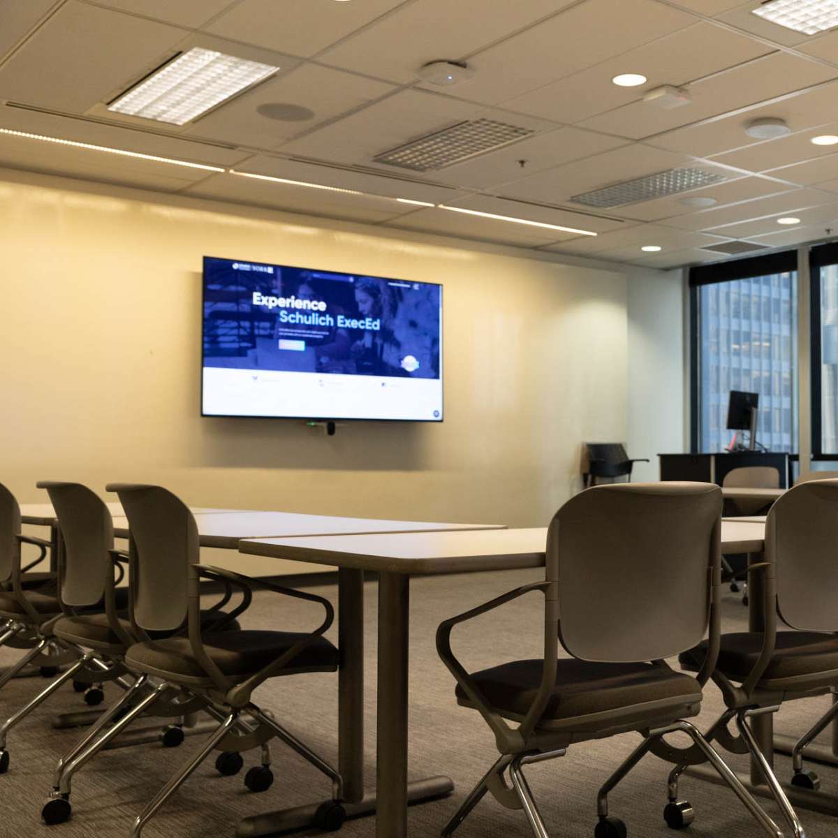 A modern conference room features a large screen on one wall, multiple tables arranged in a U-shape, and ergonomic chairs.