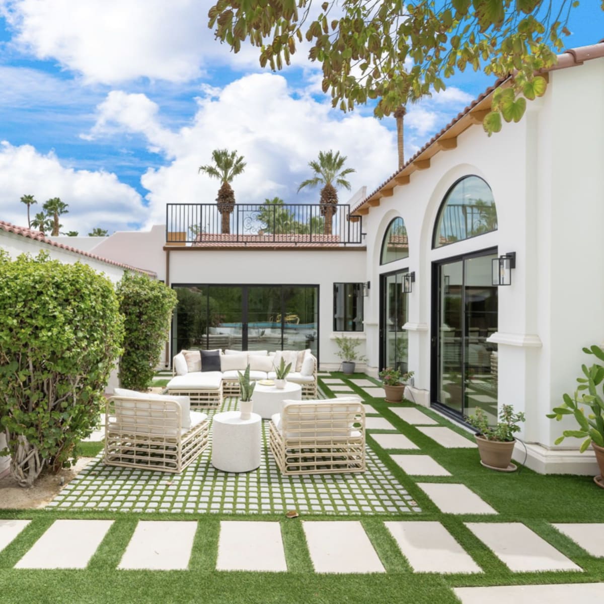 A landscaped outdoor area featuring white patio furniture, stone pathways, and lush greenery adjacent to a modern house with large windows.