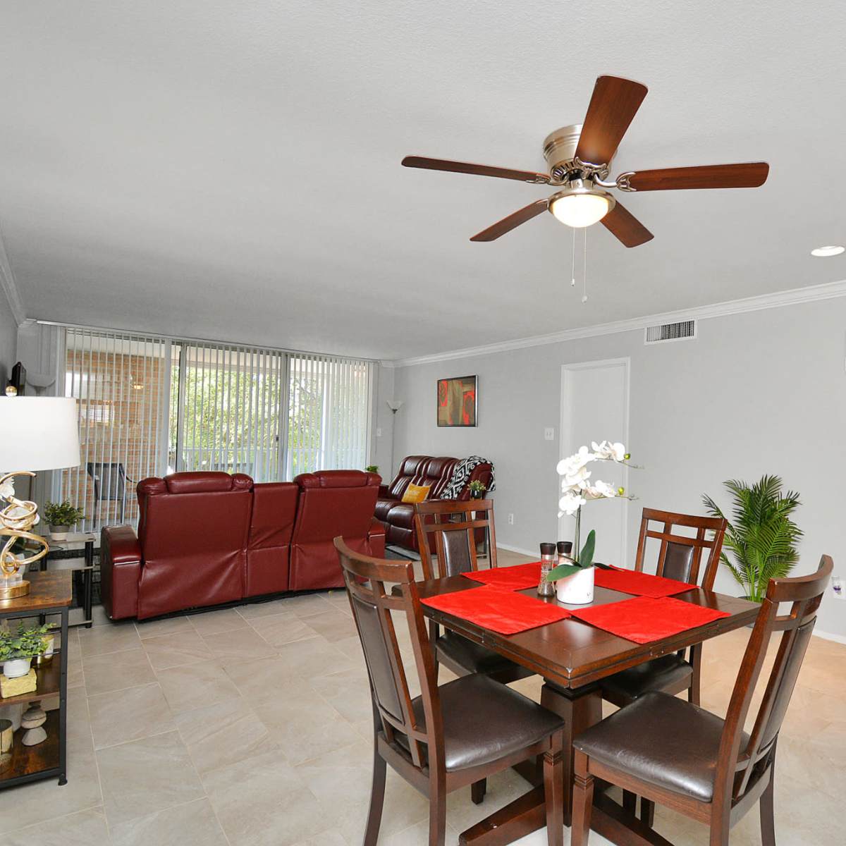 A dining area features a table with a red centerpiece, surrounded by brown chairs, with a living space visible in the background.