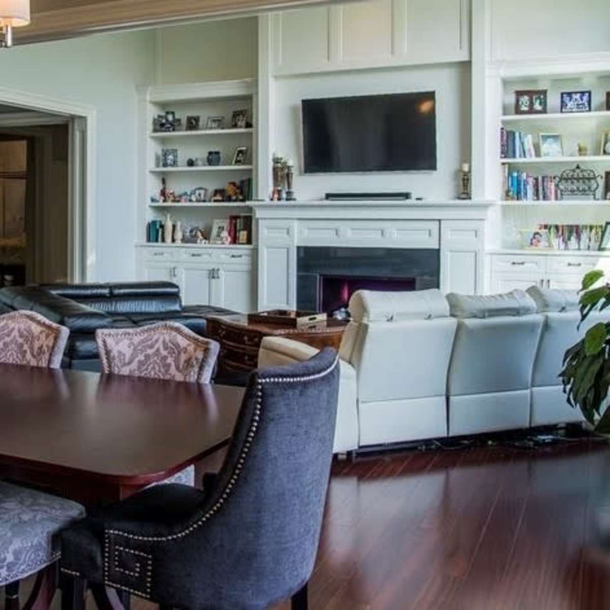 A dining area with a large wooden table surrounded by upholstered chairs, leading into a living room with a sectional sofa and a wall-mounted television.