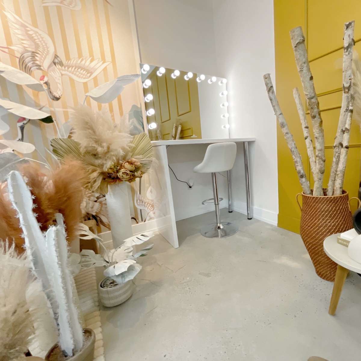 A bright room with a vanity mirror, a white chair, and various decorative plants arranged around a minimalist table.
